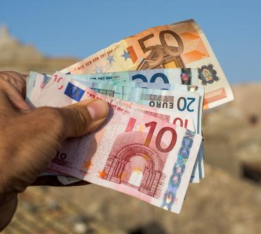 Ανδρικό χέρι κρατάει χαρτονομίσματα σε ευρώ και στο βάθος φαίνεται νησί το καλοκαίρι