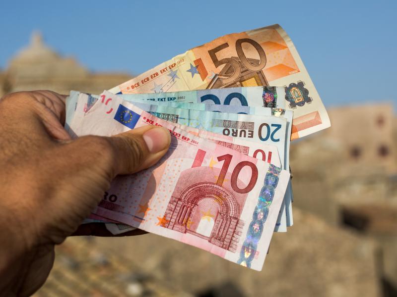 Ανδρικό χέρι κρατάει χαρτονομίσματα σε ευρώ και στο βάθος φαίνεται νησί το καλοκαίρι