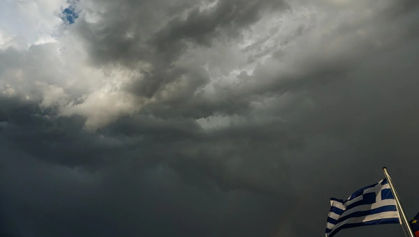 Μαύρα σύννεφα στον ουρανό πίσω από μια ελληνική σημαία