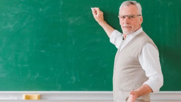 Καθηγητής δείχνει με κιμωλία σε πίνακα κοιτάζοντας προς την κάμερα