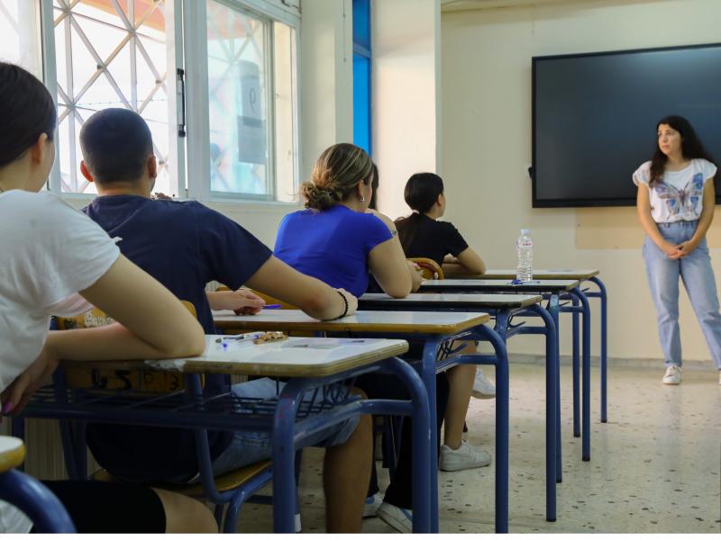 Μαθητές δίνουν πανελλήνιες, κάθονται σε θρανία και τους μιλάει δασκάλα