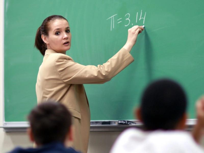 Εκπαιδευτικός σε σχολική τάξη γράφει με κιμωλία στον πράσινο πίνακα τη μαθηματική σταθερά π=3,14, ενώ παραδίδει μάθημα στους μαθητές της