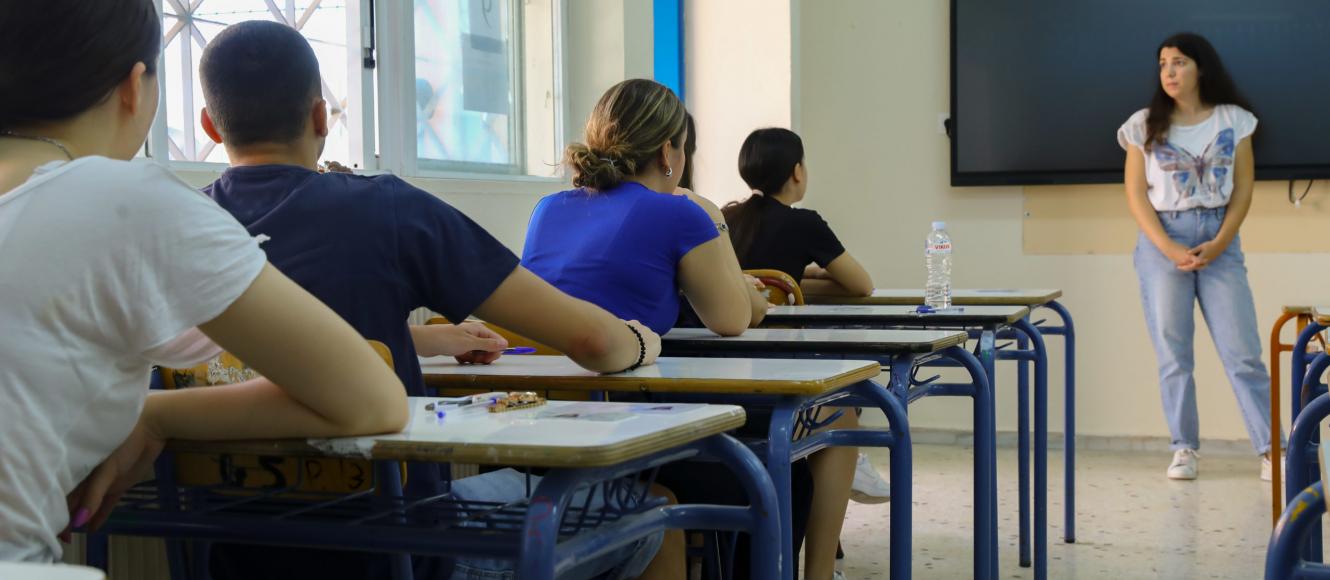 Μαθητές δίνουν πανελλήνιες, κάθονται σε θρανία και τους μιλάει δασκάλα