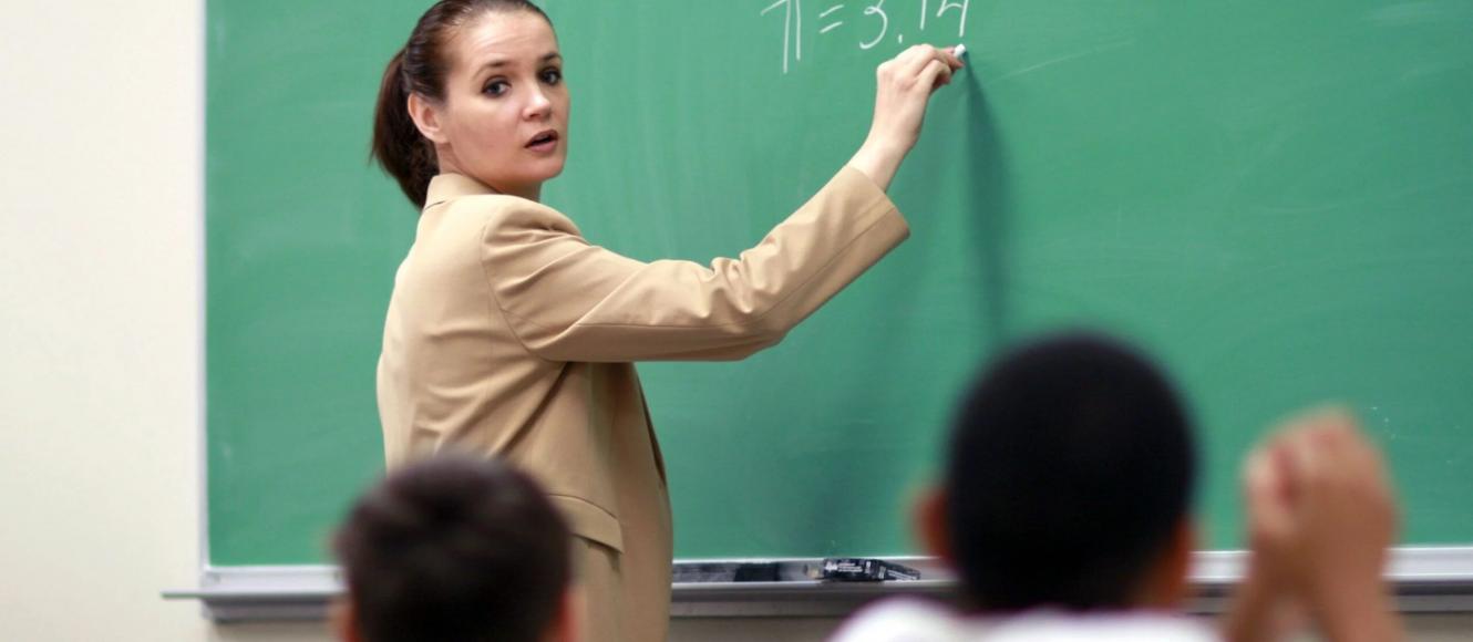 Εκπαιδευτικός σε σχολική τάξη γράφει με κιμωλία στον πράσινο πίνακα τη μαθηματική σταθερά π=3,14, ενώ παραδίδει μάθημα στους μαθητές της