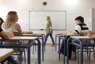 Γυναίκα εκπαιδευτικός προχωράει μέσα σε σχολική αίθουσα, στα θρανία κάθονται μαθητές