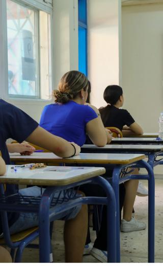 Μαθητές δίνουν πανελλήνιες, κάθονται σε θρανία και τους μιλάει δασκάλα