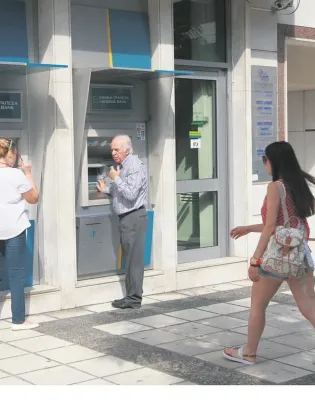 Ηλικιωμένος άνδρας και γυναίκα πραγματοποιούν συναλλαγές σε εξωτερικά ATM της Εθνικής Τράπεζας στο πεζοδρόμιο, ενώ μια κοπέλα περνάει από μπροστά.