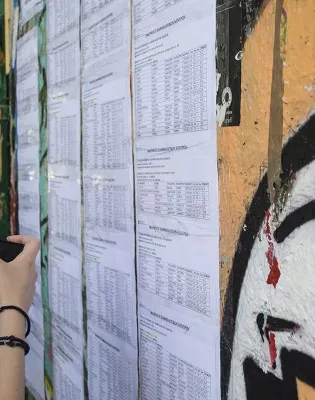 Εκτύπωση με τις βάσεις εισαγωγής των Πανελλαδικών εξετάσεων αναρτημένη σε τοίχο σχολείου, ενώ σε πρώτο πλάνο μια υποψήφια κρατάει το κινητό της τηλέφωνο