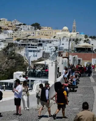 Τουρίστες περπατούν σε σοκάκι ελληνικού νησιού με φόντο παραδοσιακά λευκά σπίτια και εκκλησία με γαλάζιο τρούλο, για το πρόγραμμα Τουρισμός για Όλους