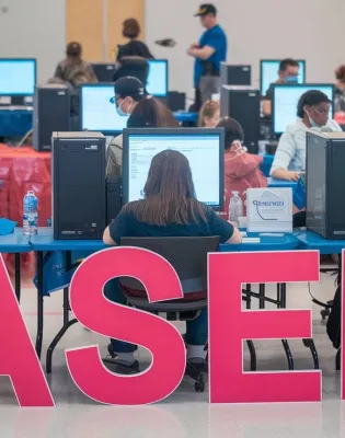 Άνθρωποι κάθονται σε κομπιούτερ με το λογότυπο του ΑΣΕΠ στα αγγλικά να βρίσκεται μπροστά τους