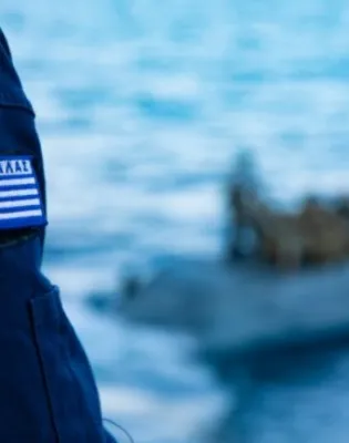 Λιμενοφύλακας με στολή Λιμενικού και ελληνική σημαία δίπλα στη θάλασσα κοιτάζει βάρκα