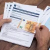 Μια κοντινή φωτογραφία, τραβηγμένη από ψηλά, που δείχνει ένα ζευγάρι τριχωτά ανδρικά χέρια να κρατούν και να χειρίζονται επίσημα έγγραφα πάνω σε ένα ξύλινο γραφείο. Το αριστερό χέρι κρατά μια στοίβα εγγράφων, με την κορυφαία σελίδα να φέρει τον τίτλο «AITHΣH ΕΠΙΣΤΡΟΦΗΣ ΕΝΟΙΚΙΟΥ»