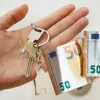 Ανθρώπινο χέρι που κρατάει κλειδιά σπιτιού με μπρελόκ και χαρτονομίσματα των 50 ευρώ, συμβολίζοντας την επιδότηση ενοικίου.