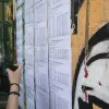 Εκτύπωση με τις βάσεις εισαγωγής των Πανελλαδικών εξετάσεων αναρτημένη σε τοίχο σχολείου, ενώ σε πρώτο πλάνο μια υποψήφια κρατάει το κινητό της τηλέφωνο