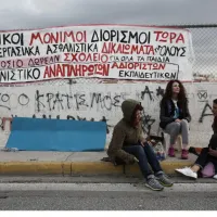 Εκπαιδευτικοί κάθονται σε πεζοδρόμιο με πανό από πίσω τους