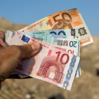 Ανδρικό χέρι κρατάει χαρτονομίσματα σε ευρώ και στο βάθος φαίνεται νησί το καλοκαίρι