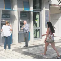 Ηλικιωμένος άνδρας και γυναίκα πραγματοποιούν συναλλαγές σε εξωτερικά ATM της Εθνικής Τράπεζας στο πεζοδρόμιο, ενώ μια κοπέλα περνάει από μπροστά.
