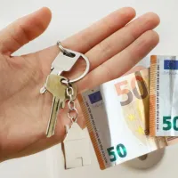 Ανθρώπινο χέρι που κρατάει κλειδιά σπιτιού με μπρελόκ και χαρτονομίσματα των 50 ευρώ, συμβολίζοντας την επιδότηση ενοικίου.