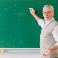 Καθηγητής δείχνει με κιμωλία σε πίνακα κοιτάζοντας προς την κάμερα