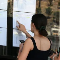 Κοντινό πλάνο δύο γυναικών που κοιτάζουν εκτυπωμένους πίνακες αποτελεσμάτων αναρτημένους σε τζάμι σχολείου. Μια νεαρή κοπέλα δείχνει με το δάχτυλό της ένα σημείο στη λίστα των βάσεων, ενώ δίπλα της μια άλλη γυναίκα παρακολουθεί προσεκτικά, κρατώντας το κινητό της τηλέφωνο