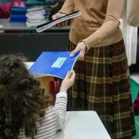 Δασκάλα δίνει μπλε τετράδιο σε μαθήτρια που κάθεται σε θρανίο