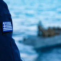 Λιμενοφύλακας με στολή Λιμενικού και ελληνική σημαία δίπλα στη θάλασσα κοιτάζει βάρκα