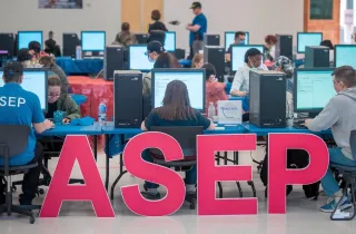 Άνθρωποι κάθονται σε κομπιούτερ με το λογότυπο του ΑΣΕΠ στα αγγλικά να βρίσκεται μπροστά τους