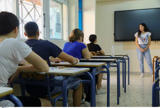 Μαθητές δίνουν πανελλήνιες, κάθονται σε θρανία και τους μιλάει δασκάλα