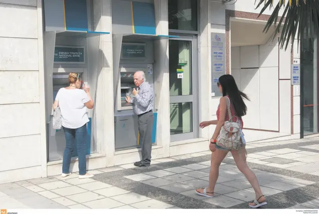 Ηλικιωμένος άνδρας και γυναίκα πραγματοποιούν συναλλαγές σε εξωτερικά ATM της Εθνικής Τράπεζας στο πεζοδρόμιο, ενώ μια κοπέλα περνάει από μπροστά.