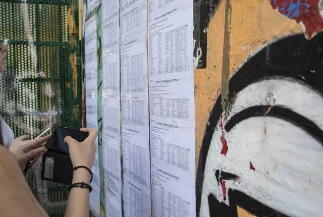 Εκτύπωση με τις βάσεις εισαγωγής των Πανελλαδικών εξετάσεων αναρτημένη σε τοίχο σχολείου, ενώ σε πρώτο πλάνο μια υποψήφια κρατάει το κινητό της τηλέφωνο