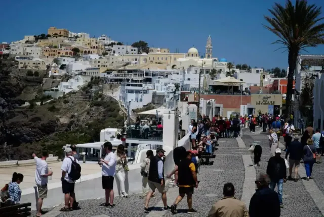 Τουρίστες περπατούν σε σοκάκι ελληνικού νησιού με φόντο παραδοσιακά λευκά σπίτια και εκκλησία με γαλάζιο τρούλο, για το πρόγραμμα Τουρισμός για Όλους