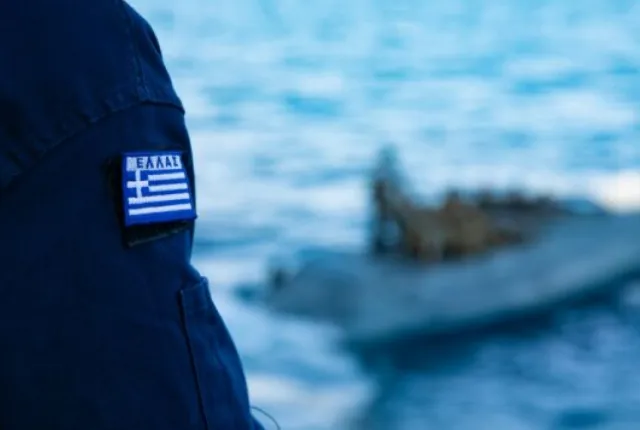 Λιμενοφύλακας με στολή Λιμενικού και ελληνική σημαία δίπλα στη θάλασσα κοιτάζει βάρκα