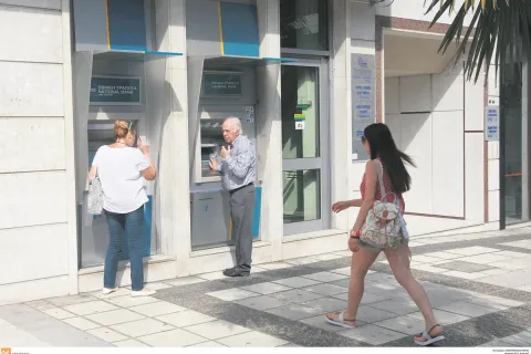 Ηλικιωμένος άνδρας και γυναίκα πραγματοποιούν συναλλαγές σε εξωτερικά ATM της Εθνικής Τράπεζας στο πεζοδρόμιο, ενώ μια κοπέλα περνάει από μπροστά.