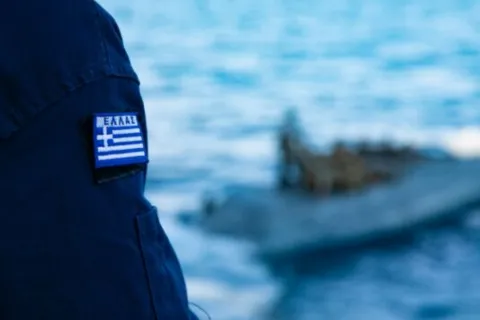 Λιμενοφύλακας με στολή Λιμενικού και ελληνική σημαία δίπλα στη θάλασσα κοιτάζει βάρκα