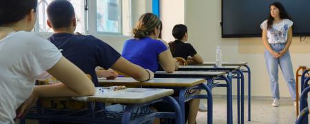 Μαθητές δίνουν πανελλήνιες, κάθονται σε θρανία και τους μιλάει δασκάλα